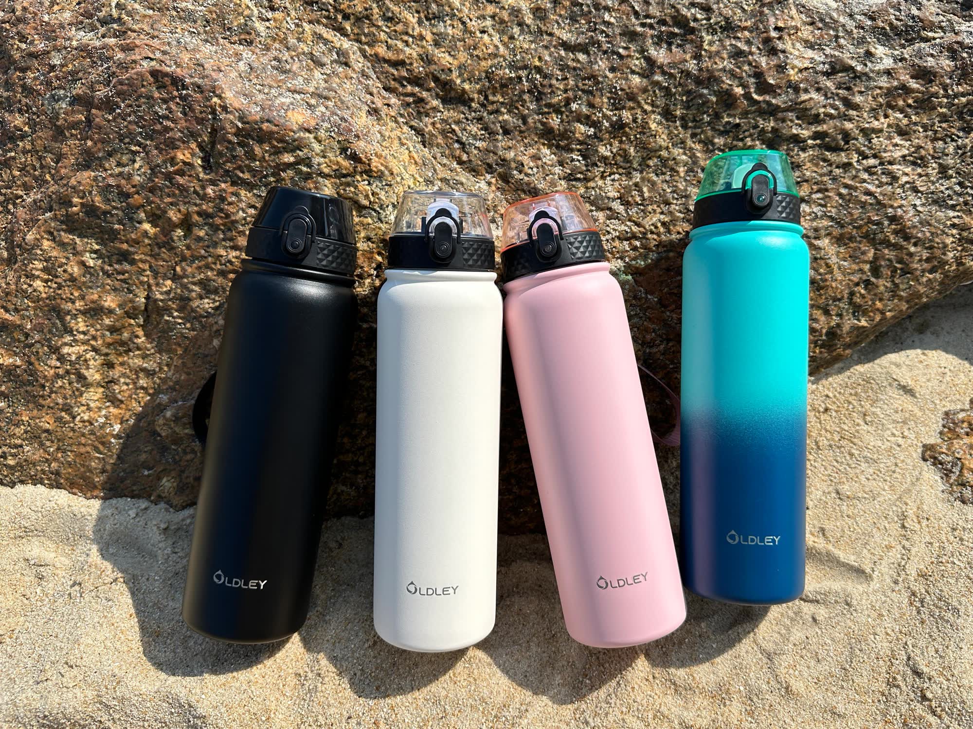 four solid color water bottles in black white pink and teal gradient on sand near rocks