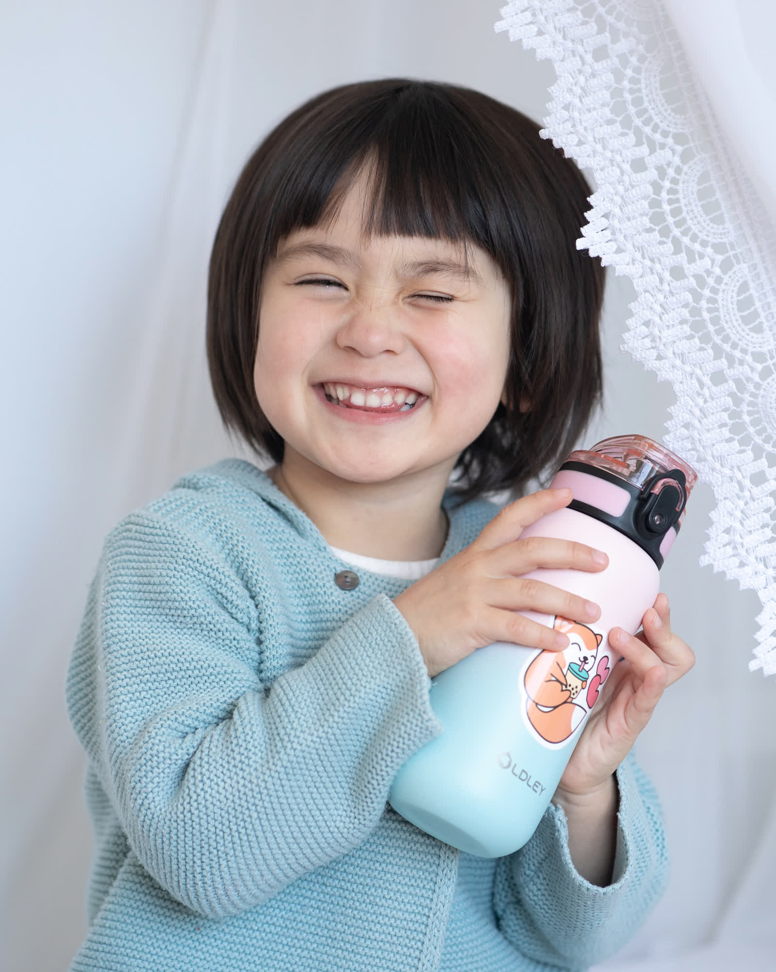 smiling young girl with short bangs wearing light blue knitted cardigan holding pastel gradient water bottle with cartoon shiba inu sticker indoors near lace curtain