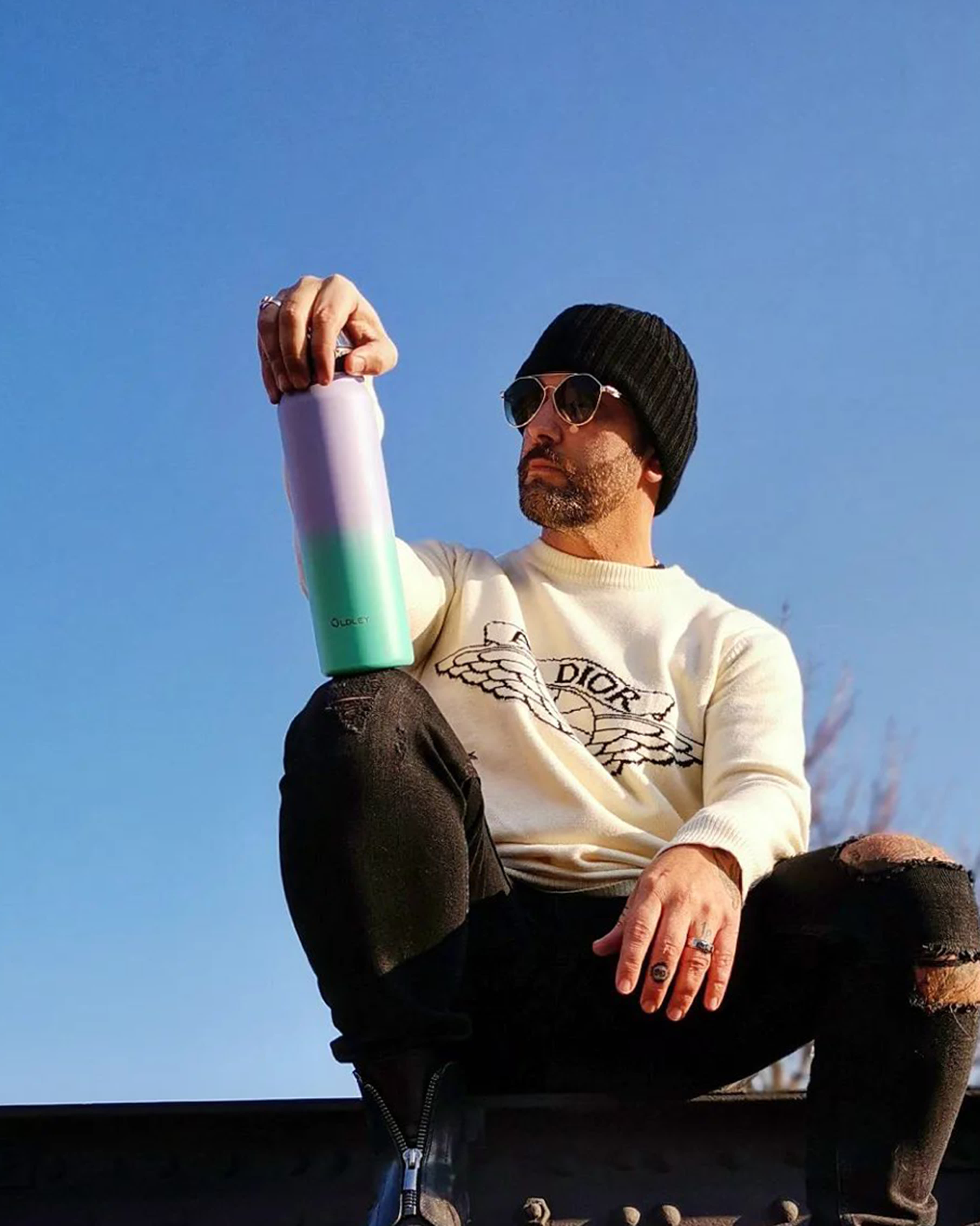 man with beard wearing black beanie and white sweatshirt holding gradient bottle against blue sky
