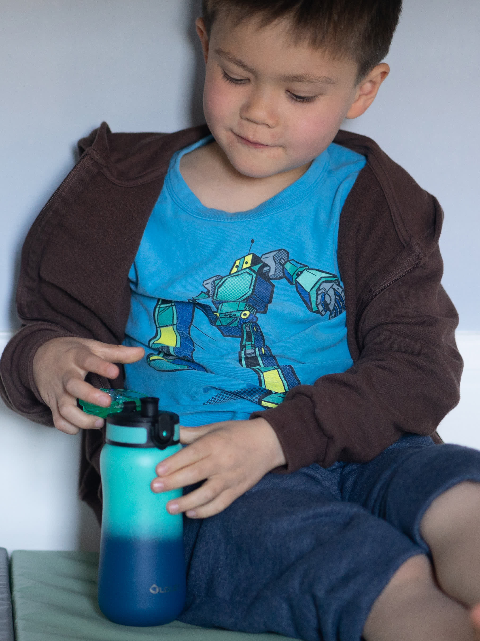 young boy wearing blue robot t shirt and brown hoodie holding teal to blue gradient water bottle while sitting indoors