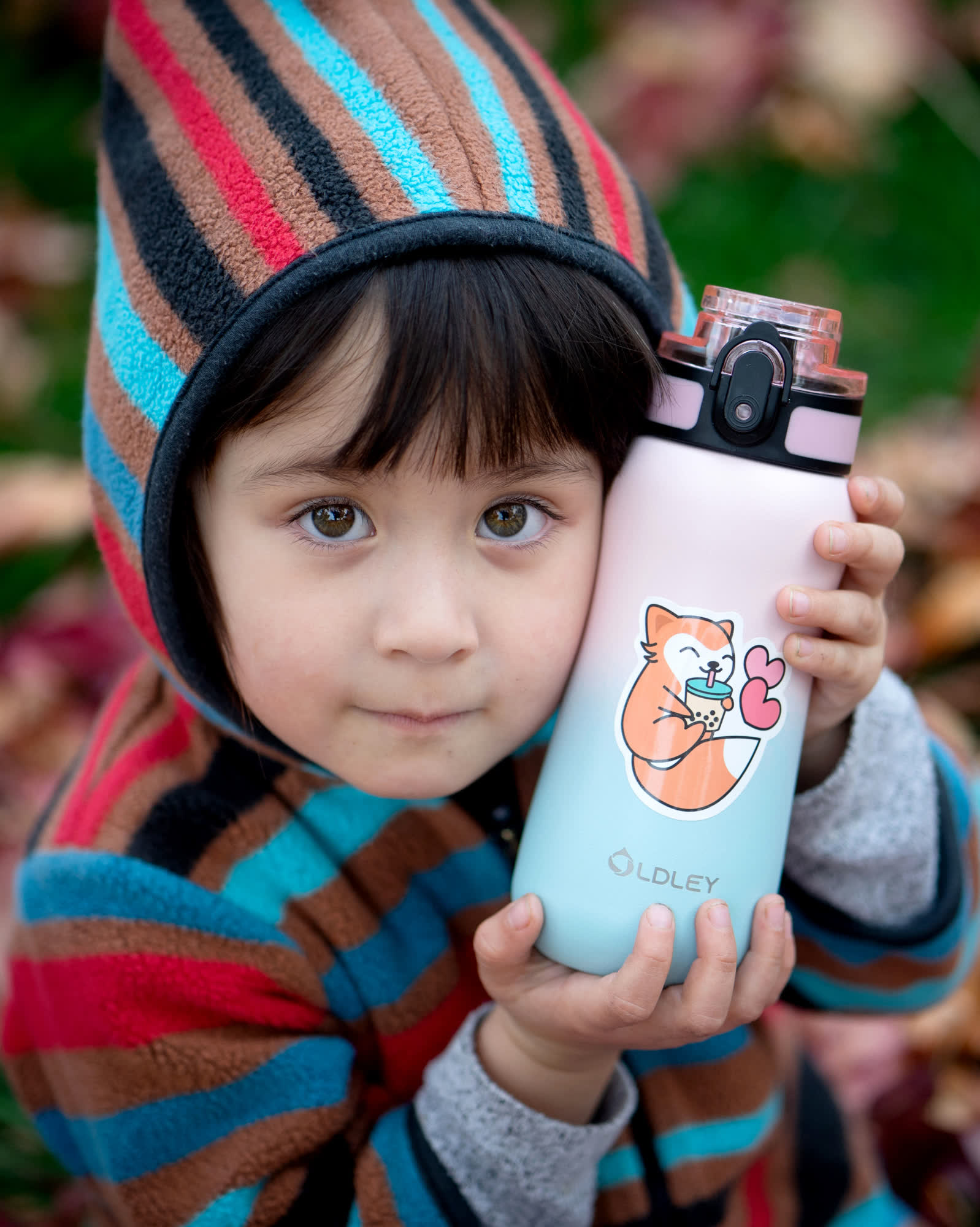 young child wearing colorful striped hooded fleece jacket holding pastel gradient water bottle with cartoon shiba inu sticker outdoors among autumn leaves