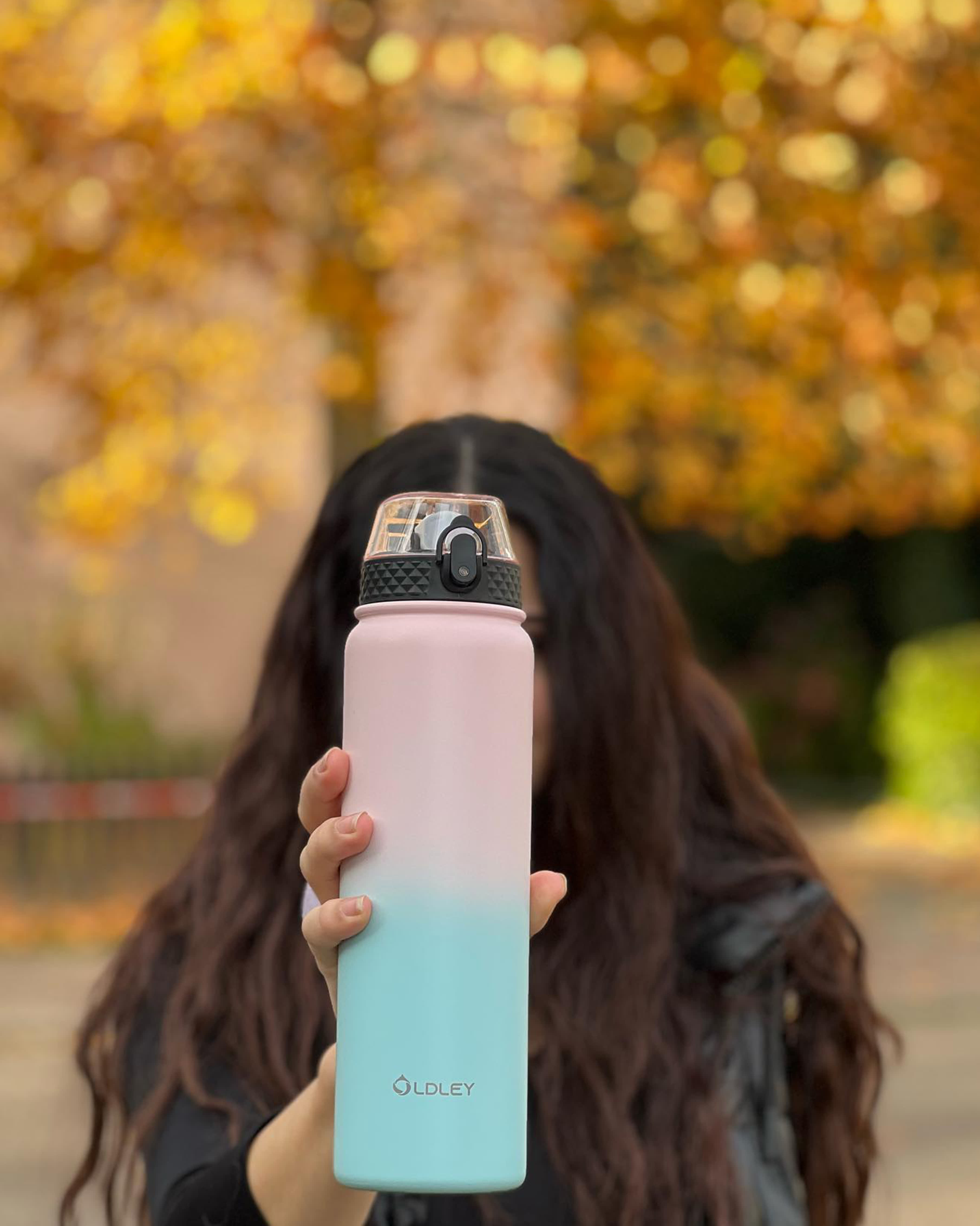 woman with long dark curly hair holding pink to blue gradient bottle with autumn leaves background