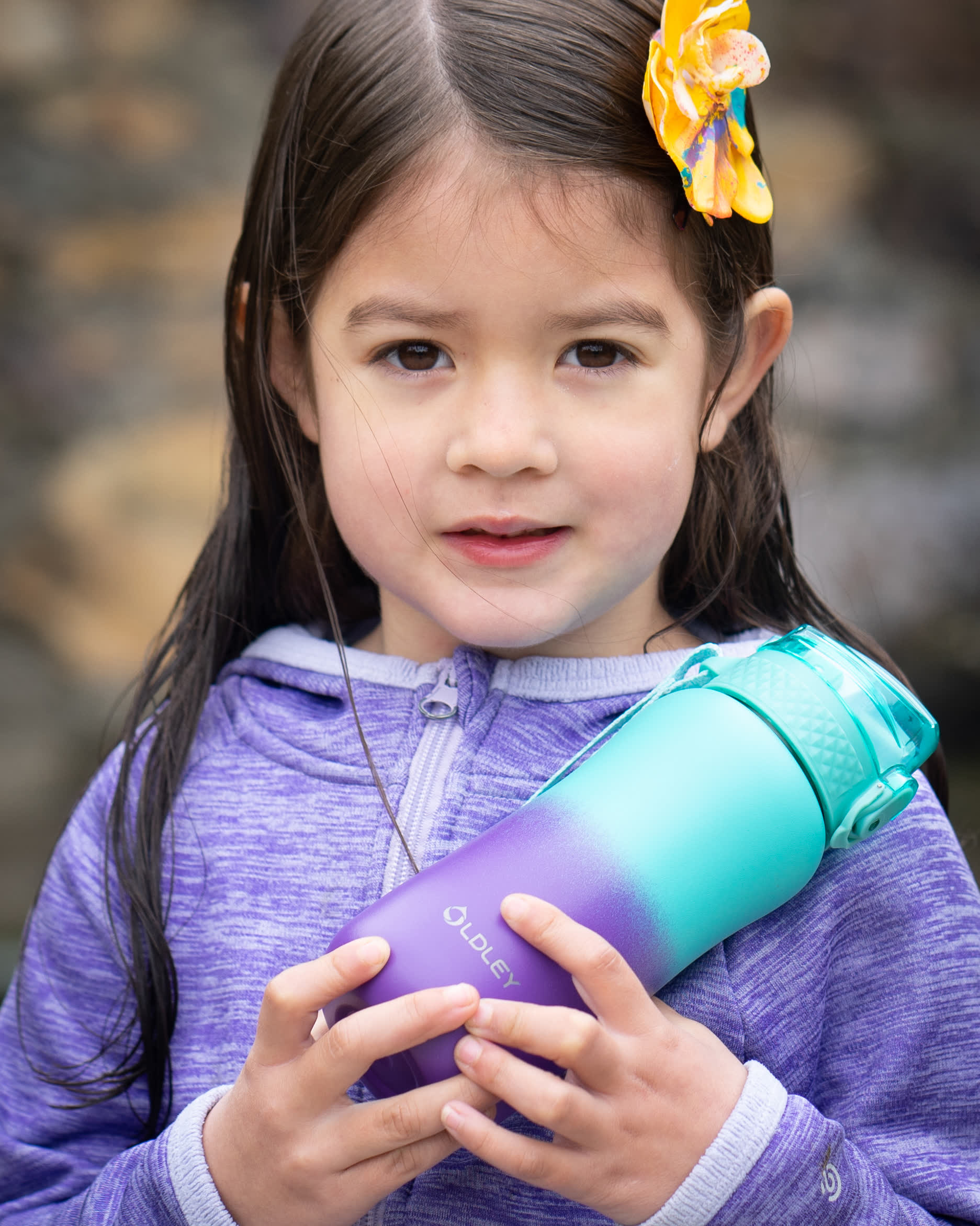 young girl with yellow flower in hair wearing purple hoodie holding gradient purple to teal insulated water bottle outdoors