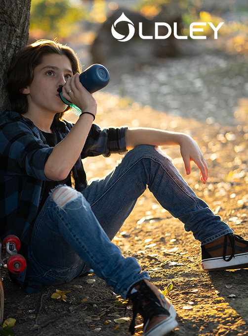 teenager in plaid shirt sitting under tree drinking from blue bottle with skateboard