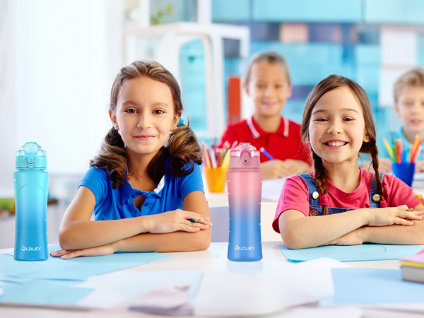 two girls sitting at school desk with blue and pink gradient water bottles and boy in red shirt in background