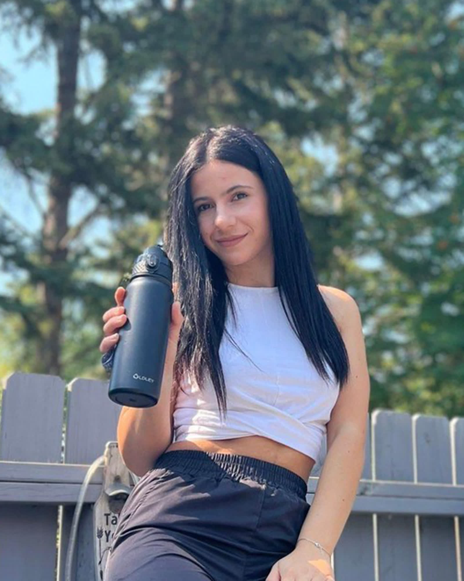 woman with long dark hair in white crop top holding navy bottle outdoors with trees and fence