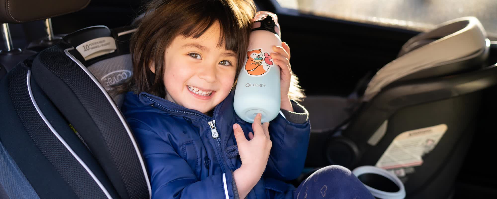 smiling child in blue puffy jacket sitting in graco car seat holding mint green water bottle with cartoon shiba inu sticker inside car