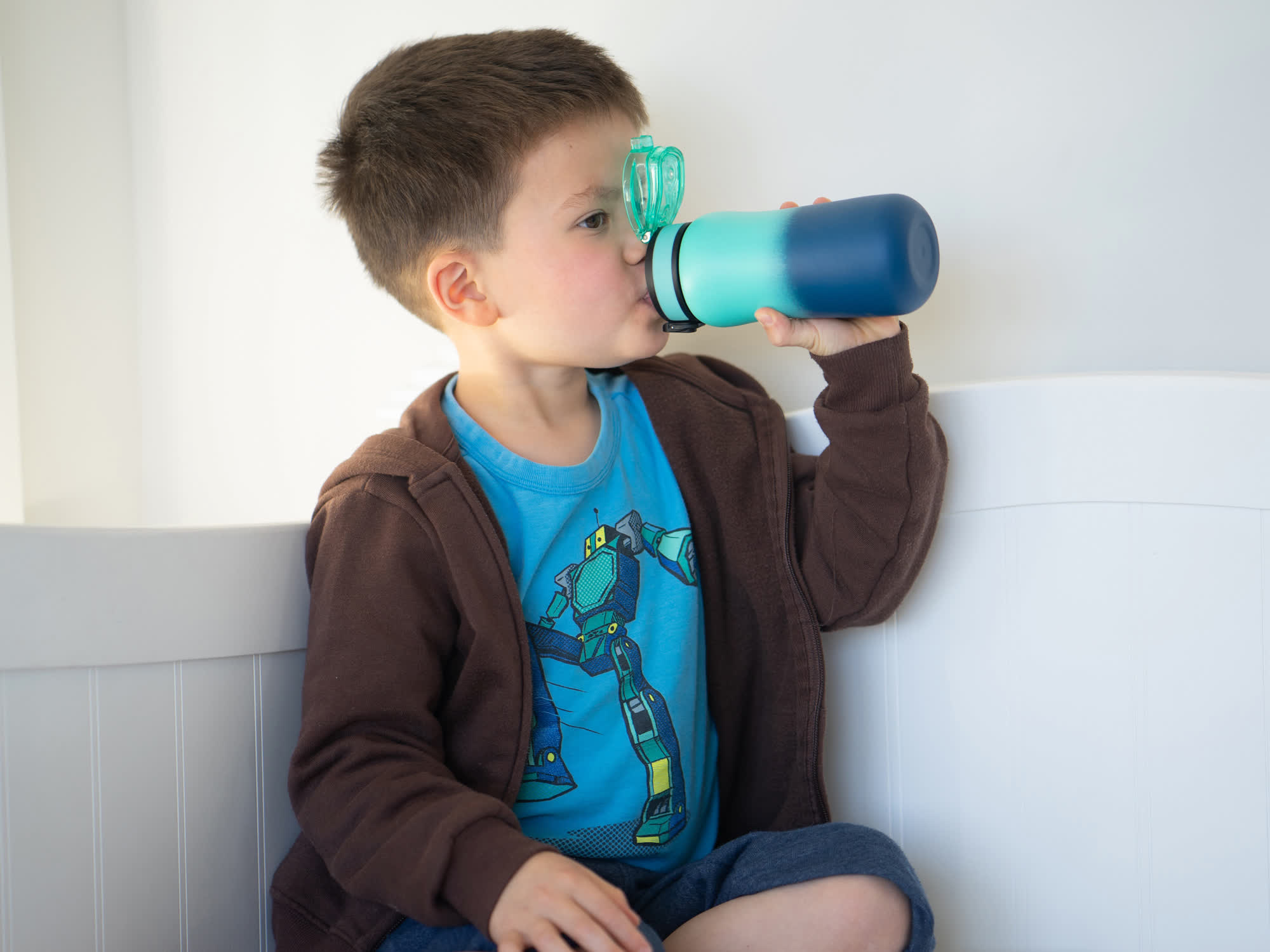 young boy in robot shirt drinking from blue water bottle against white wall